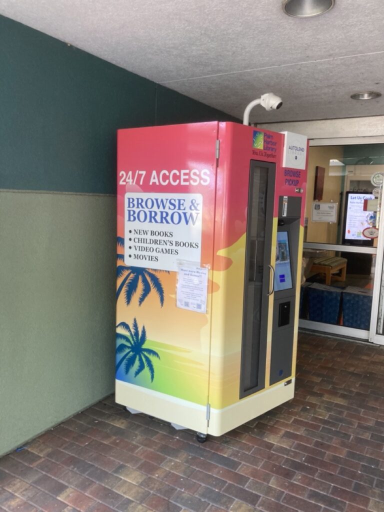 the outdoor 24/7 library borrowing kiosk outside palm harbor library. the kiosk is sunset colors with palm trees and a sign explaining the borrowing.