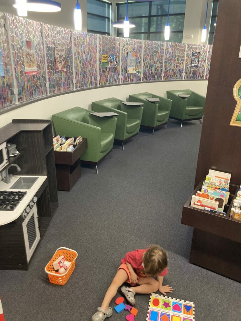 countryside children's library play area with a toddler looking down doing a shape puzzle and a play kitchen and chairs in the background