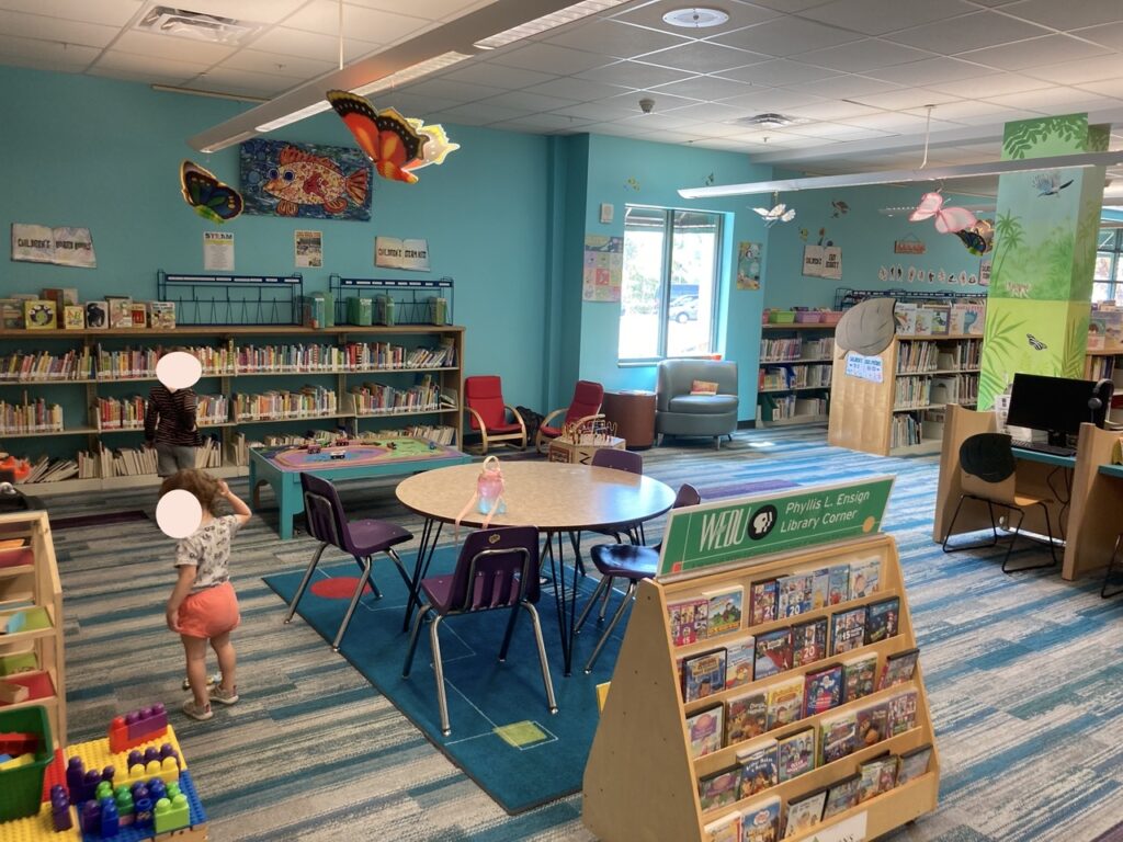 children's library area showing two toddlers with their faces obscured, a kids tables, shelves of books and toys