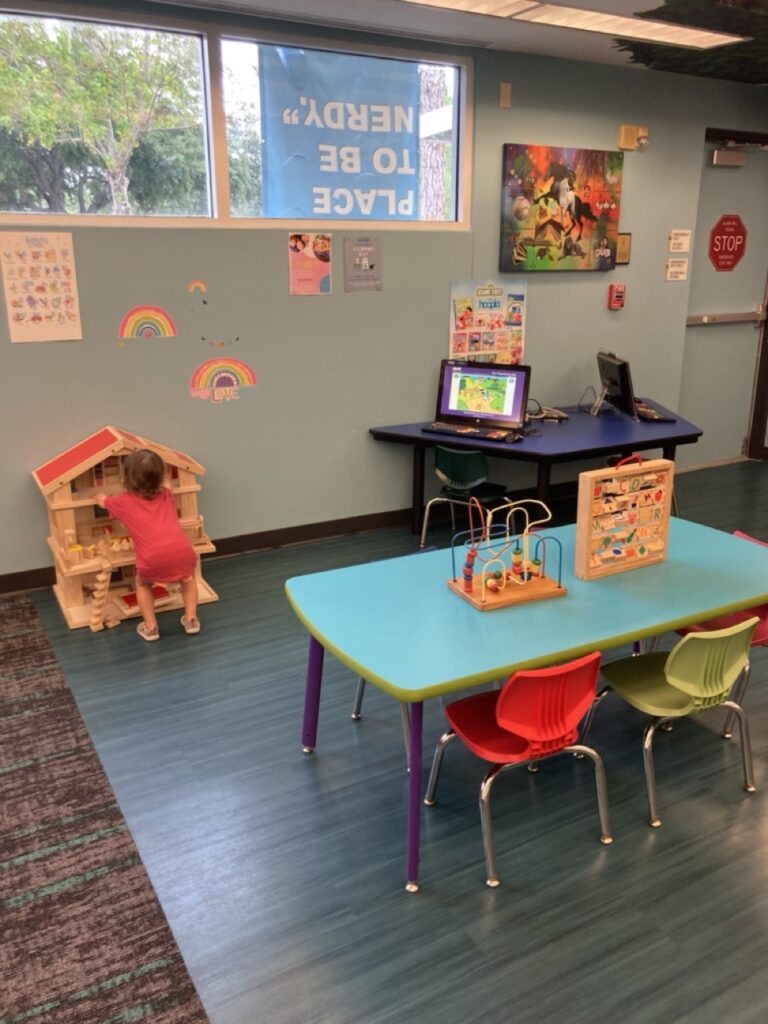 play are at palm harbor library showing two tables with toys on it and the back of a baby playing with a doll house