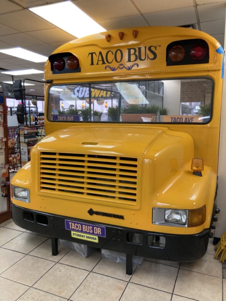 The front of a Taco Bus restaurant, it is the front of a school bus inside a building that says Taco Bus