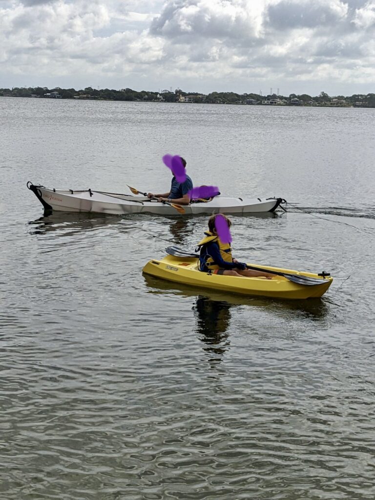 A child in a yellow small kayak and an adult in a big white kayak. Their faces are blurred out. 