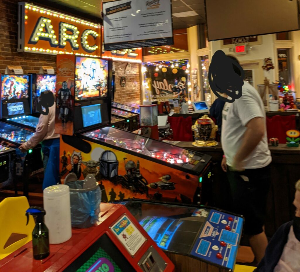 A person standing at a pinball game (with their face blacked out for privacy)
