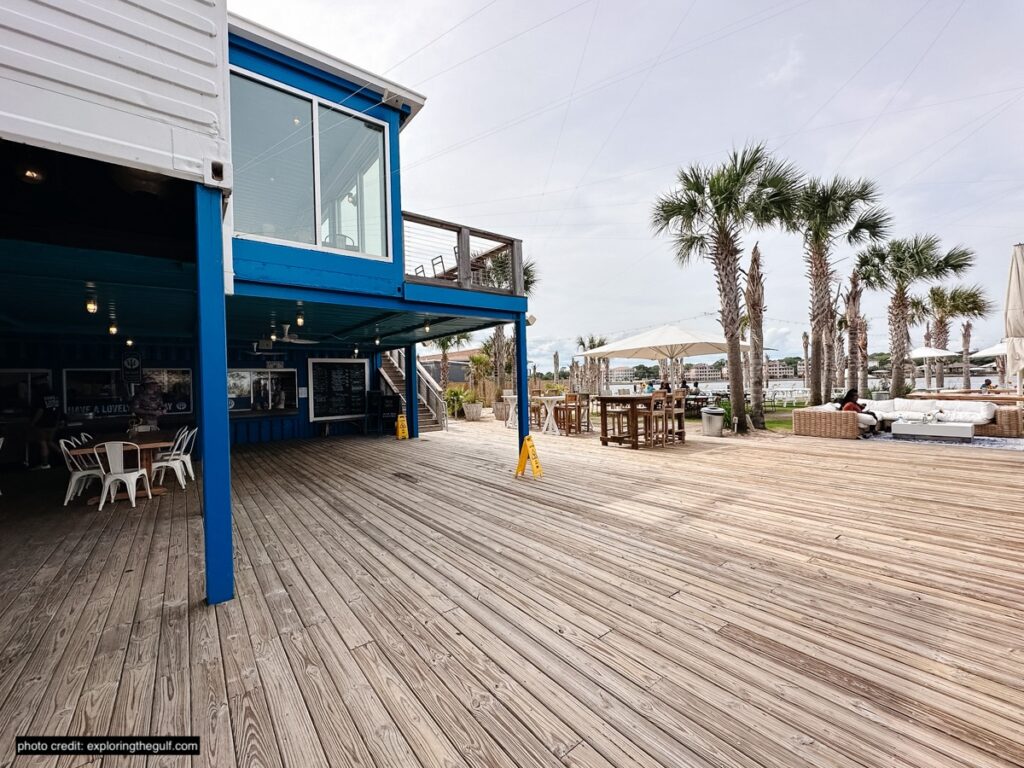 this is a picture of a wooden pier with a building in the foreground and palm trees and beach chairs in the background in destin fl