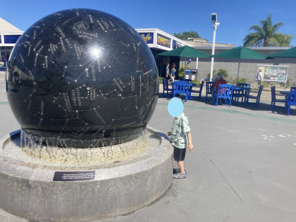 a toddler with face obscured standing in from of the spinning ball water fountain sculpture at Kennedy Space Center