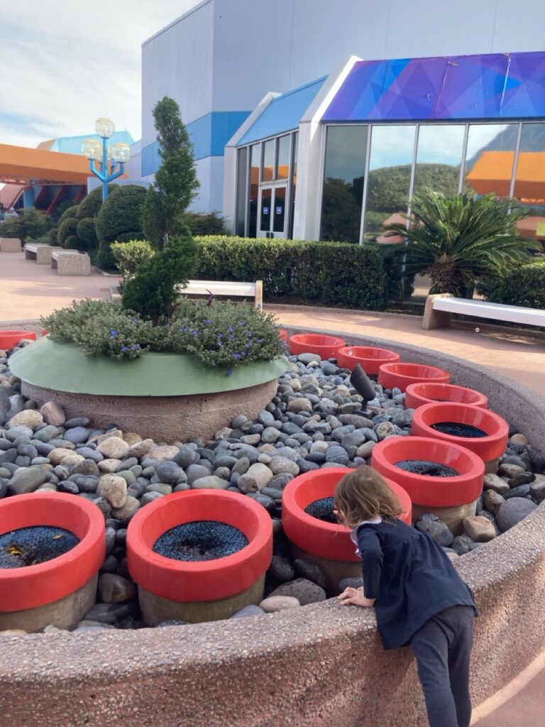 toddler leaning over to touch a fountain coming out of little red planters at epcot