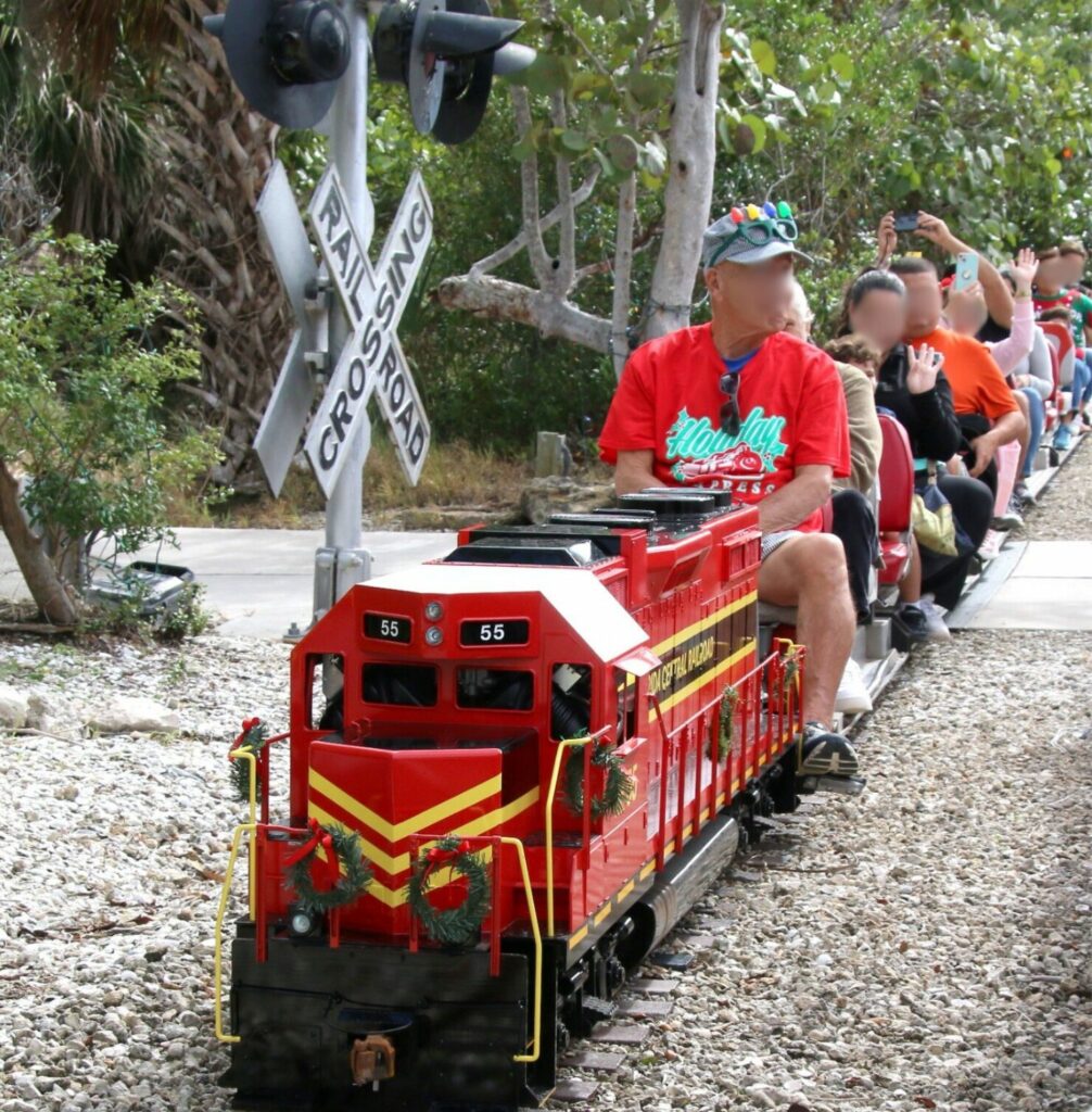 A 7.5 gauge miniature train with people riding it. The engine is red and going by a railroad crossing sign. 