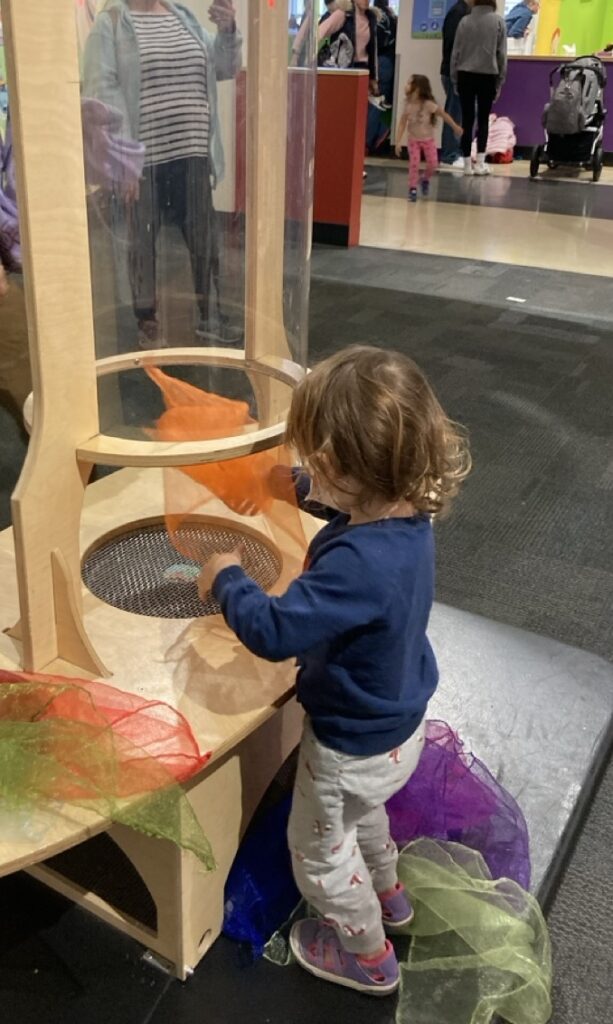 toddler playing with an orange scarf in a wind tunnel at a children's musem