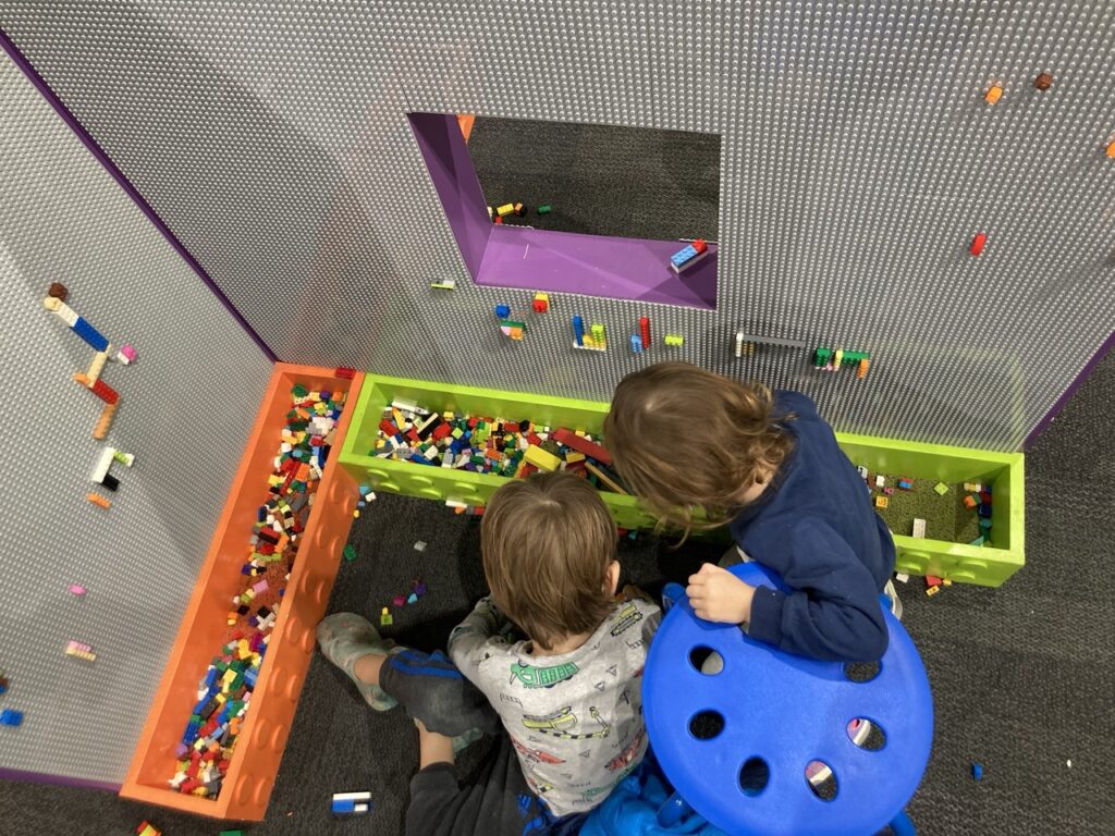 this is a photo looking down at the head of two toddlers as they look through lego bins and a lego wall next to them