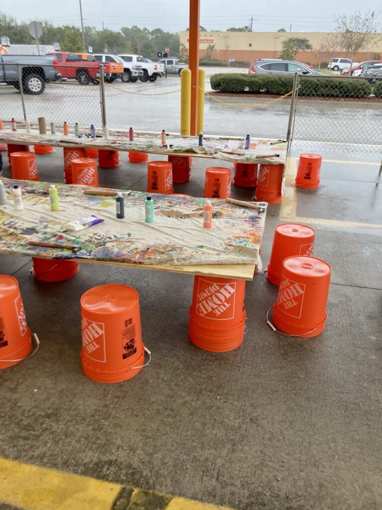 Tables made of large pieces of wood on top of orange home depot buckets, with upside down home depot buckets are chairs. There are paint for kids on the tables