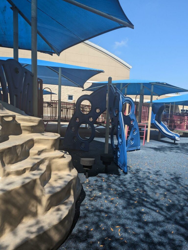 playground with blue shade canopy and plastic rock climbing area in the foreground at dunedin community center