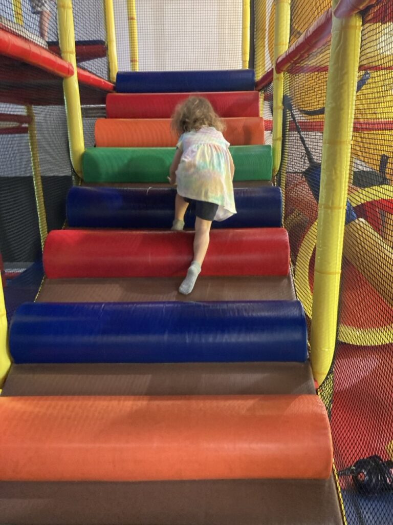 toddler climbing up a rainbow colored ramp with soft steps in an indoor playgorund