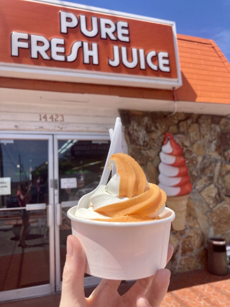Hand holding up a cup of orange-vanilla soft serve in front of a building with the sign that says Pure Fresh Juice at Yellow Banks Grove in Largo FL