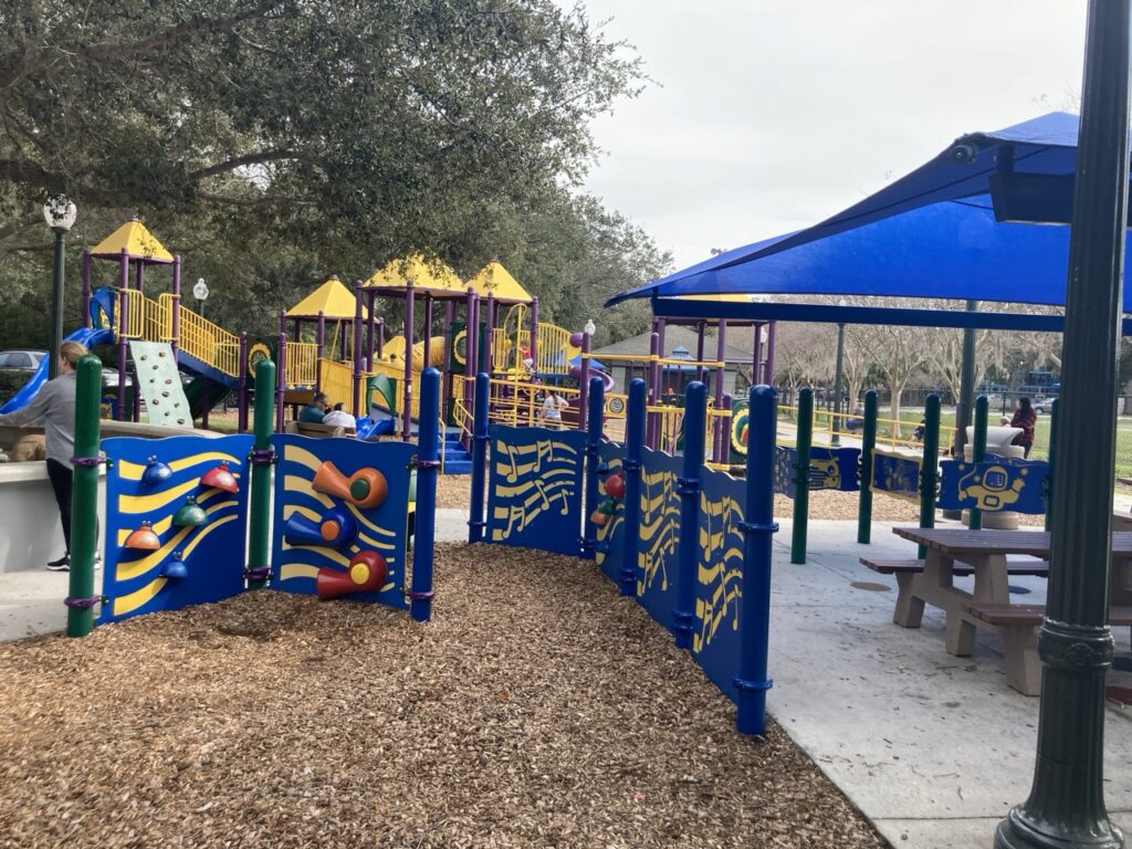 picture of playground shade canopy over picnic tables, musical instrument playground equipment and playground in the background at sunshine limitless park in clearwater